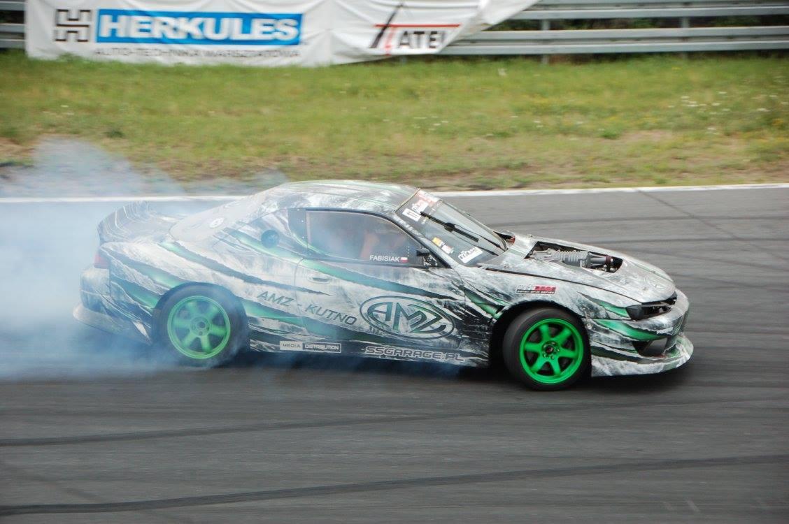 Sportowy samochód w trakcie driftu, z zielonymi felgami i niestandardowym oklejeniem w odcieniach szarości i zieleni, unoszący się dym z opon na torze.