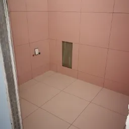 Ściany i podłoga wyłożone różowymi płytkami w trakcie prac wykończeniowych łazienki, widoczna puszka instalacyjna i otwór w ścianie.