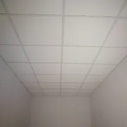 Zastosowanie sufitu podwieszanego z płyt karton gips w biurze, widoczne równe kwadratowe płyty i delikatne łączenia, w tle fragment drabiny.