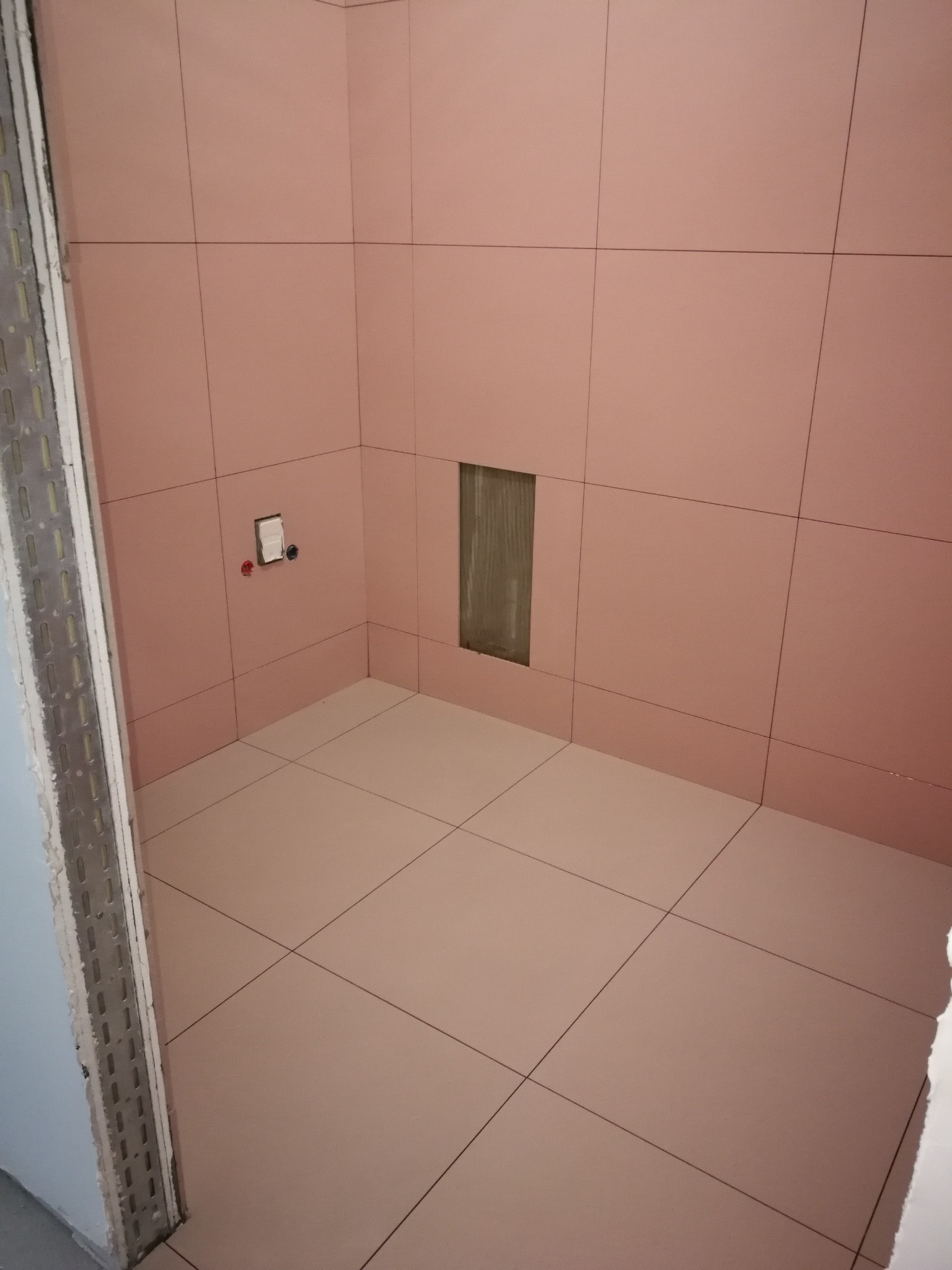 Ściany i podłoga w łazience wyłożone różowymi płytkami ceramicznymi, widoczny otwór instalacyjny w ścianie i fragment ościeżnicy drzwiowej.