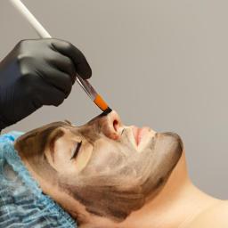 Studio Nefretete - Nakładanie czarnej maski na twarz kobiety pędzlem, pacjentka w czepku ochronnym, widok z boku.