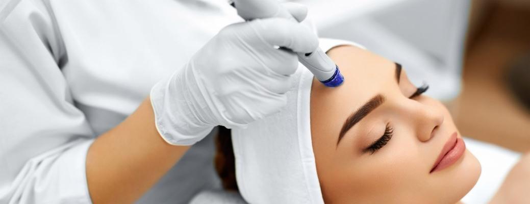 Zabieg hydrafacial na czole kobiety z zamkniętymi oczami, widoczna biała rękawica i końcówka urządzenia z niebieskim płynem.