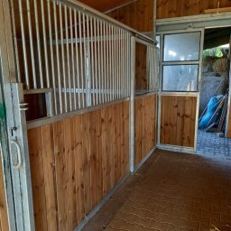 Metfach - Wnętrze stajni z metalową konstrukcją boksów i drewnianym wypełnieniem. Widoczne przesuwne elementy i wyjście na zewnątrz.