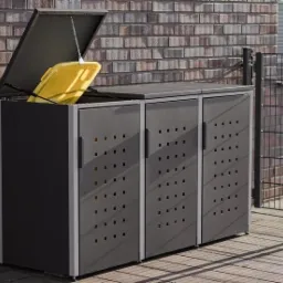 Metalowa zabudowa na trzy pojemniki na odpady, jeden z pojemników jest żółty i widoczny, z otwartą klapą, na tle ceglanego muru i metalowego ogrodzenia.