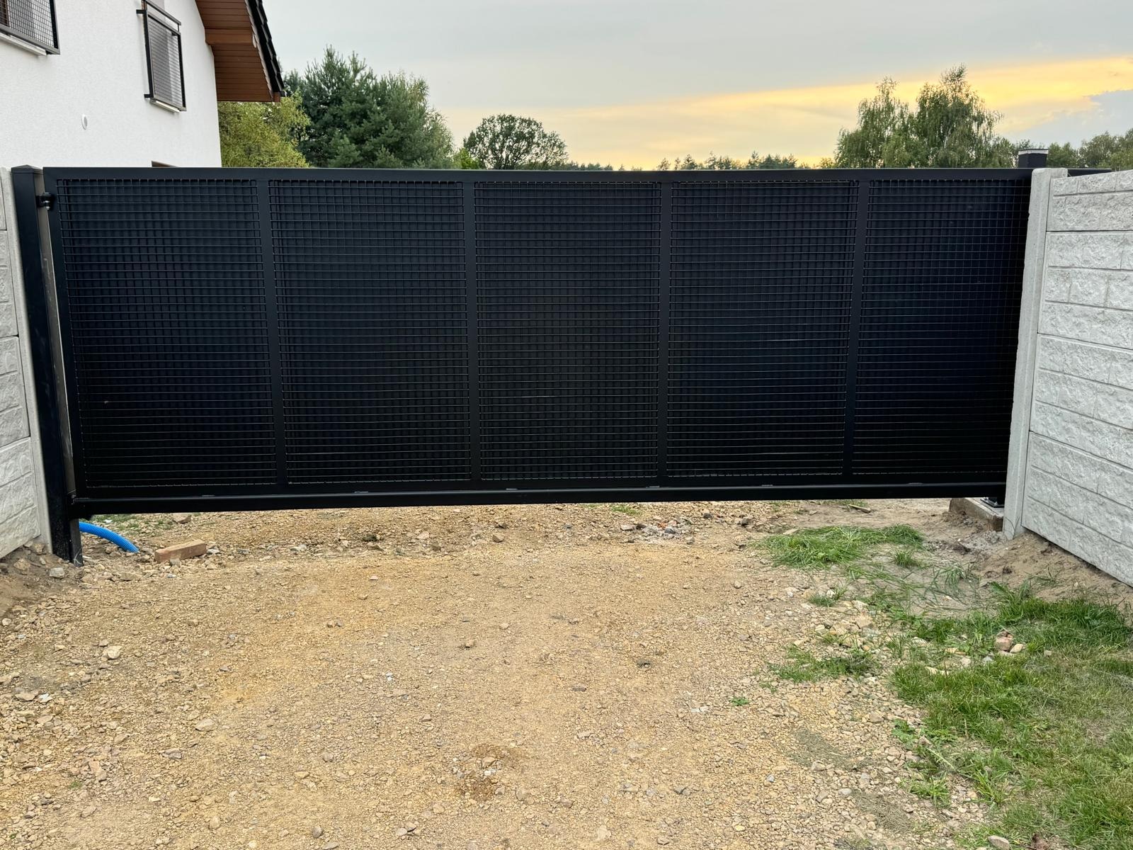 Nowoczesna, przesuwna brama w kolorze czarnym z siatkowym wypełnieniem, zamontowana na posesji. Widoczne betonowe słupki i fragment domu w tle.