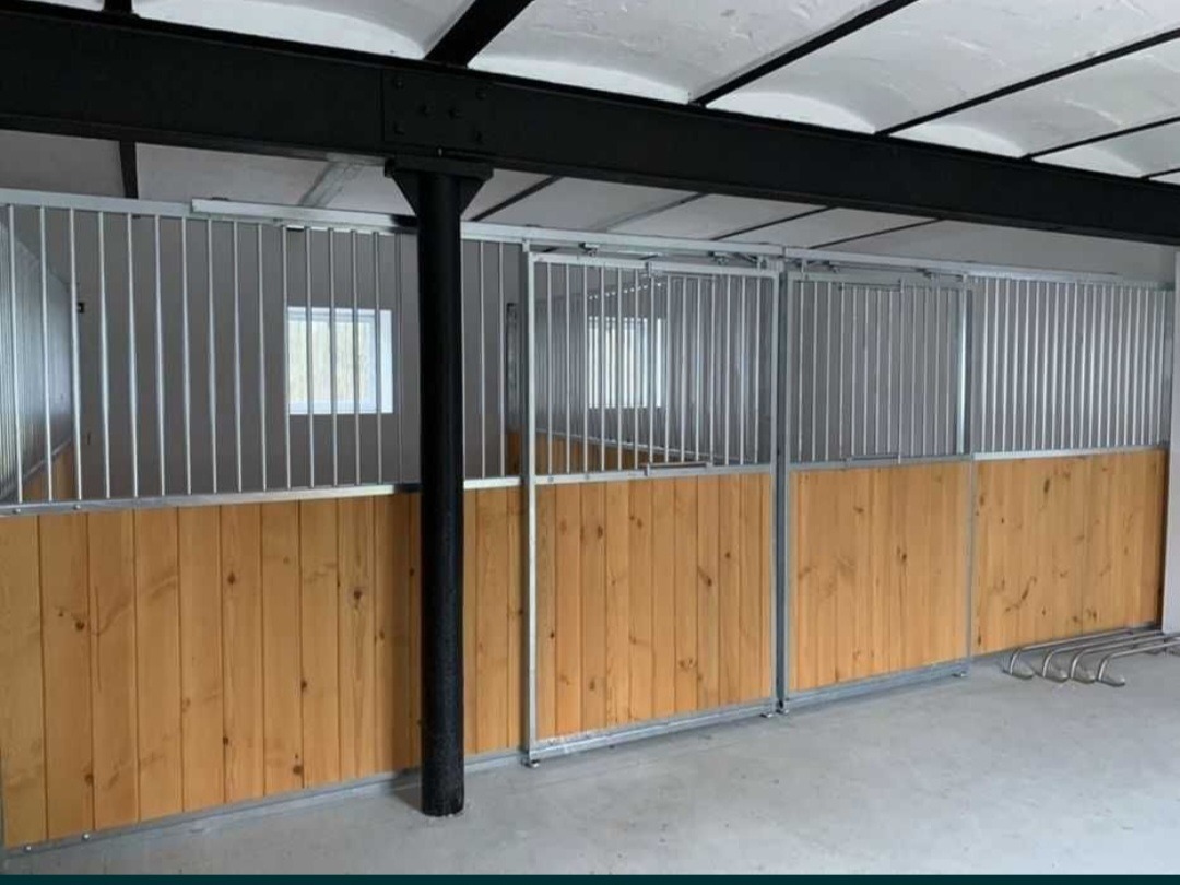 Wnętrze stajni z boksami o konstrukcji metalowo-drewnianej. Widoczna metalowa konstrukcja i drewniane panele. W tle okno z widokiem na zewnątrz.