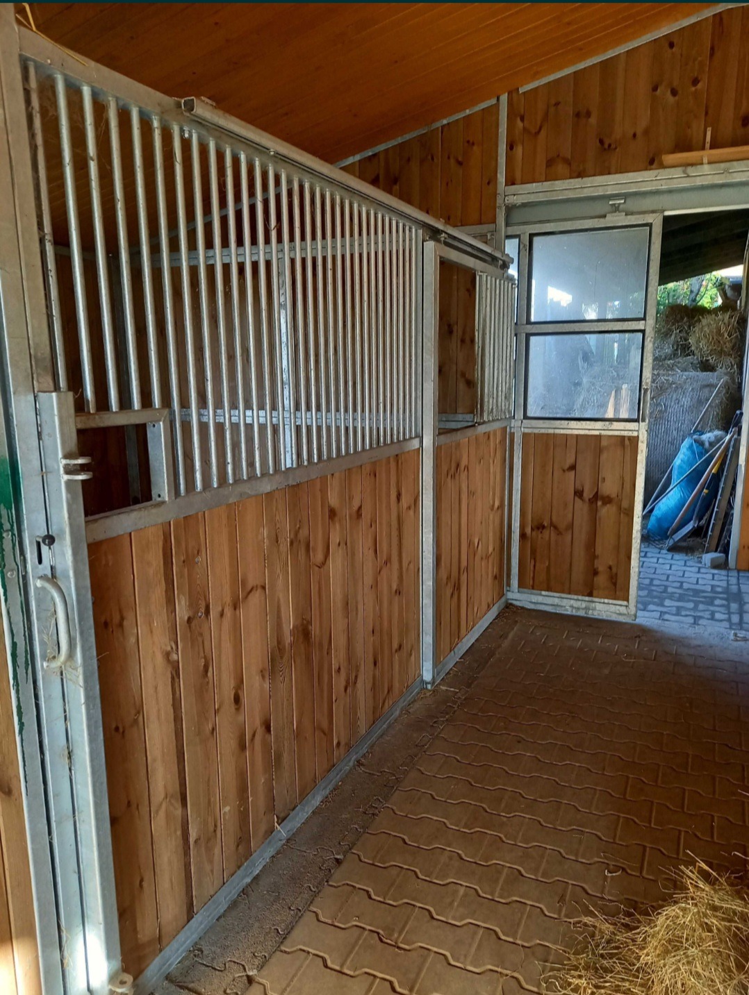 Wnętrze stajni z metalową konstrukcją boksów i drewnianym wypełnieniem. Widoczne przesuwne elementy i wyjście na zewnątrz.