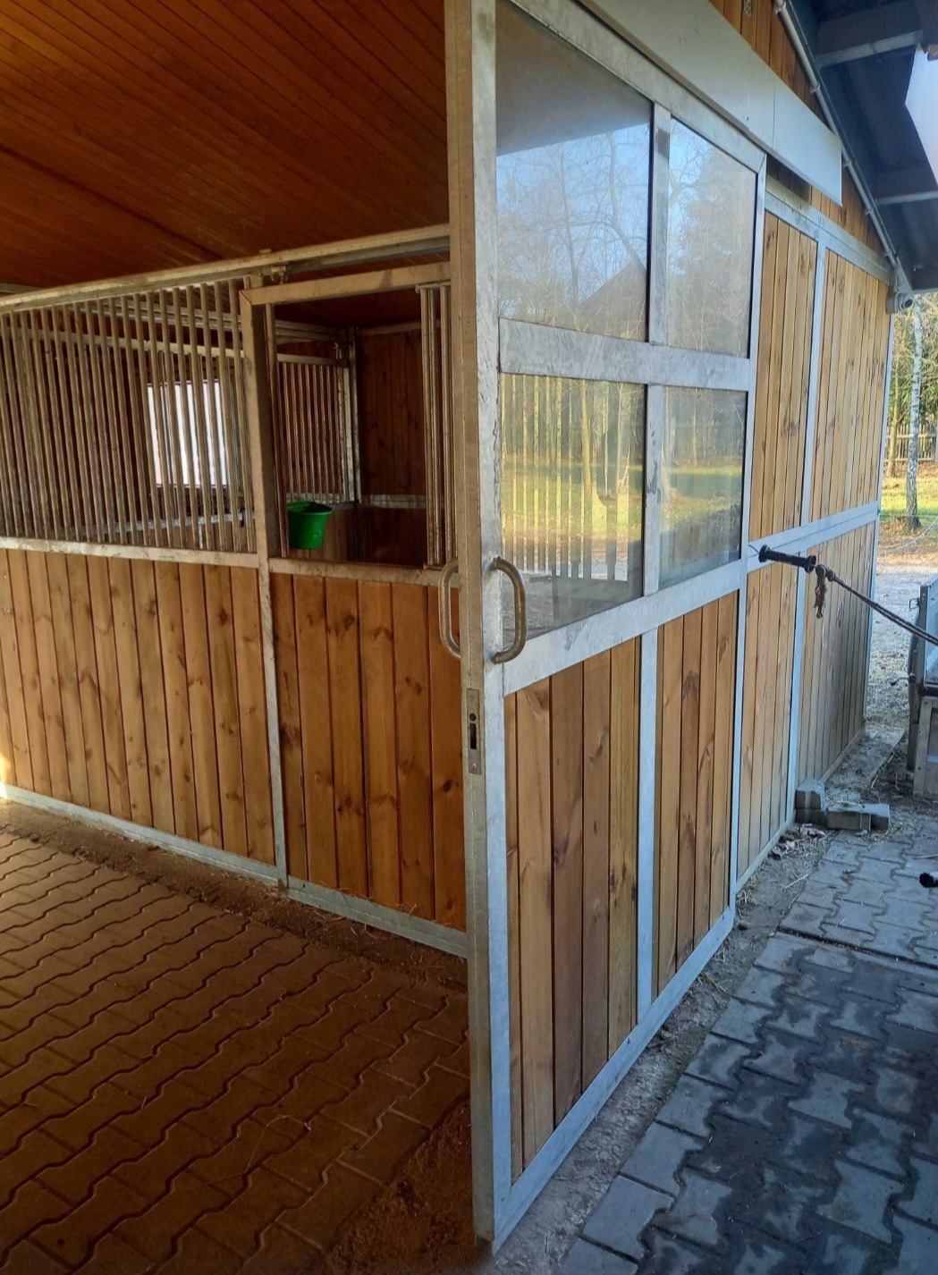 Wykończony boks dla koni z drewnianą boazerią i metalową konstrukcją. Częściowo otwarte przesuwne drzwi z szybami. Widoczna kostka brukowa i fragment otoczenia.