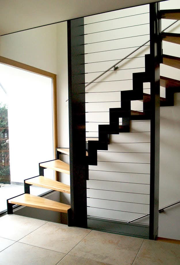 Nowoczesne schody z drewnianymi stopniami i czarną metalową konstrukcją, zabezpieczone poziomymi linkami. Widok z dołu, jasne wnętrze.