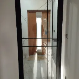 Nowoczesne drzwi wewnętrzne z czarną metalową ramą i szklanym wypełnieniem, widok na korytarz z drewnianymi drzwiami i dywanikiem.