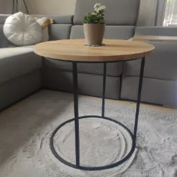 Okrągły stolik kawowy z jasnym drewnianym blatem i czarną metalową podstawą, na puszystym, jasnym dywanie, z rośliną w doniczce na blacie, ustawiony w salonie z szarą sofą.