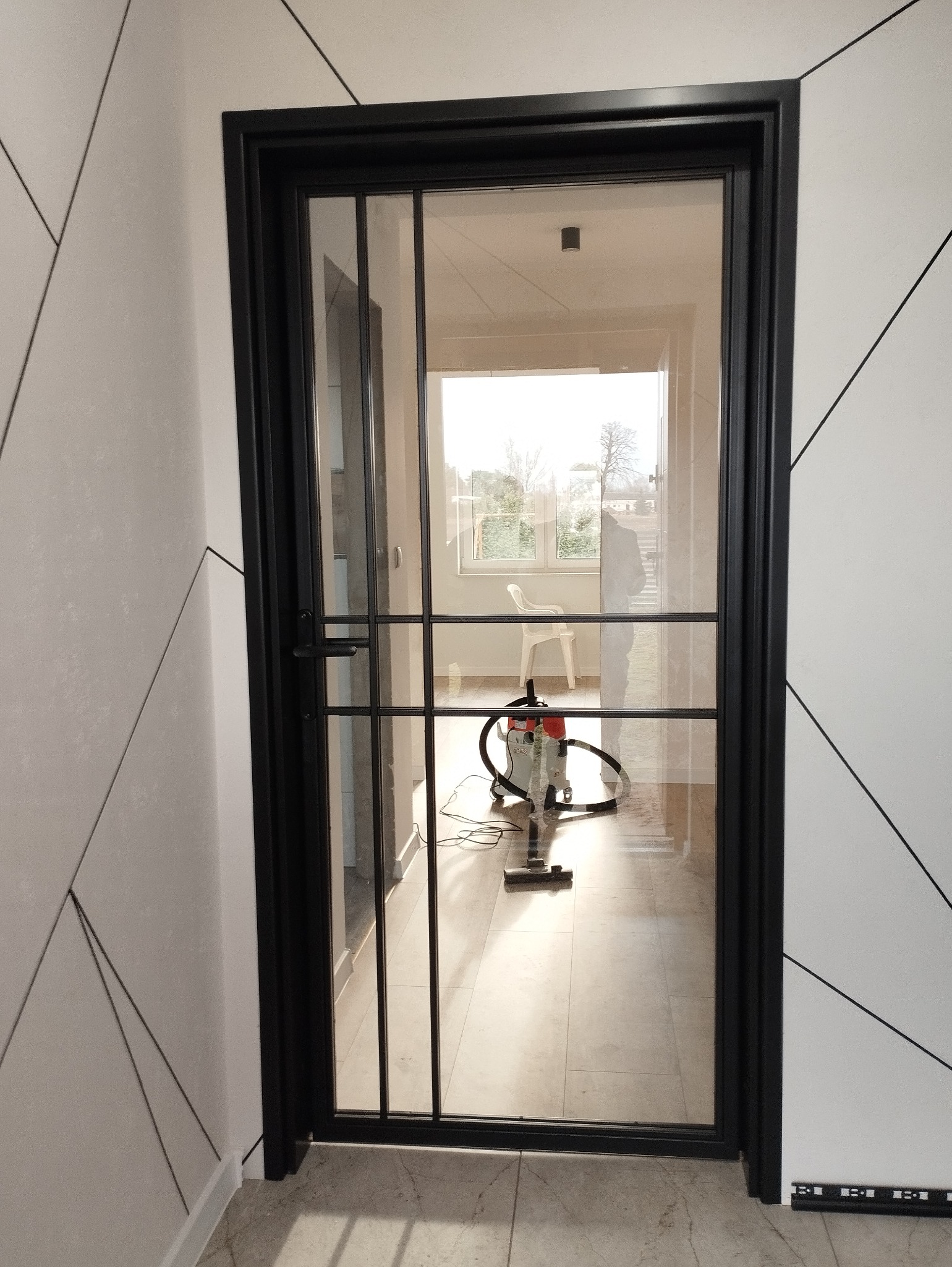 Czarna, przeszklona, metalowa konstrukcja drzwi z podziałami na tle jasnego wnętrza z odkurzaczem i białym krzesłem w tle.