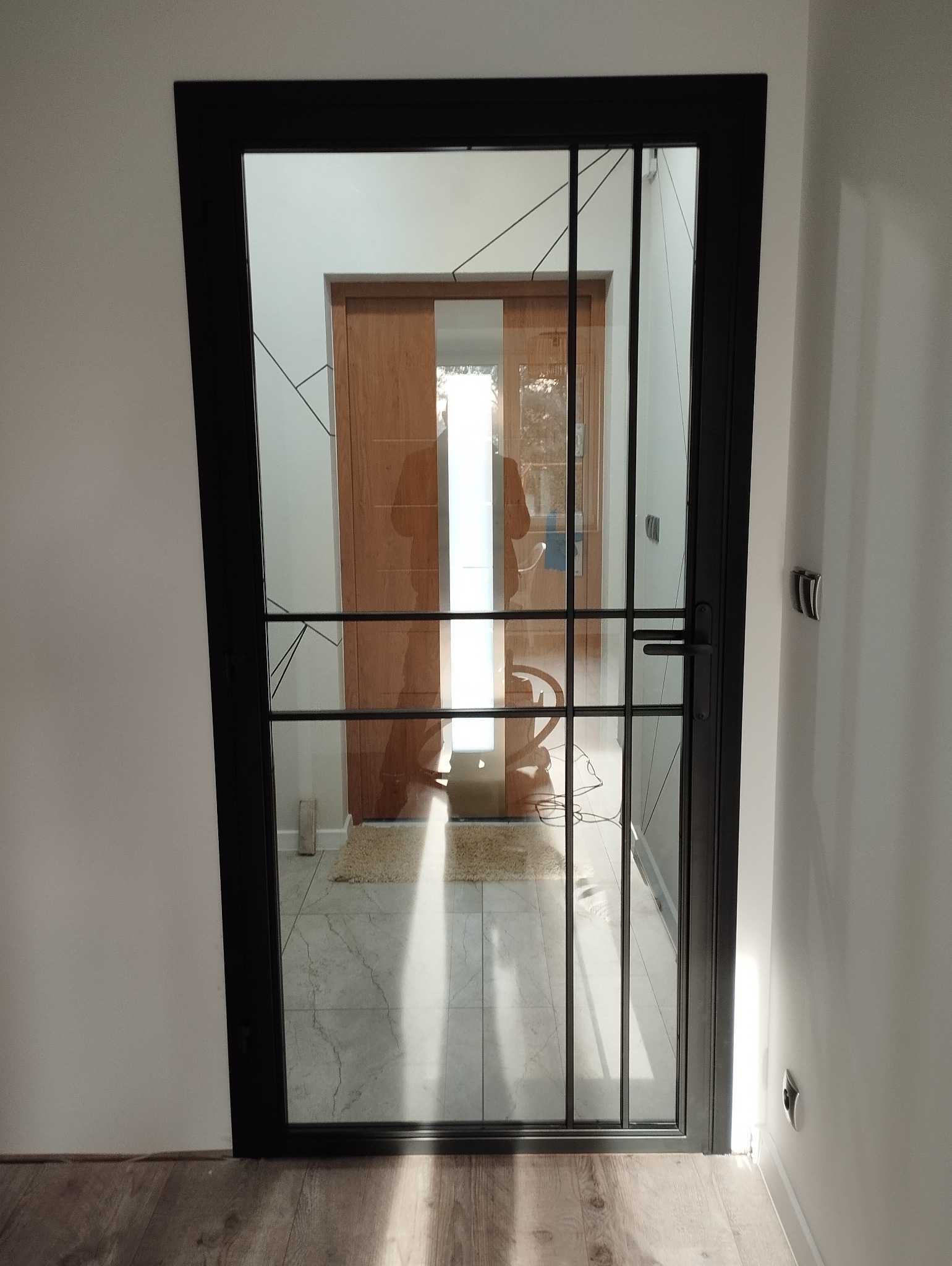 Nowoczesne drzwi wewnętrzne z czarną metalową ramą i szklanym wypełnieniem, widok na korytarz z drewnianymi drzwiami i dywanikiem.