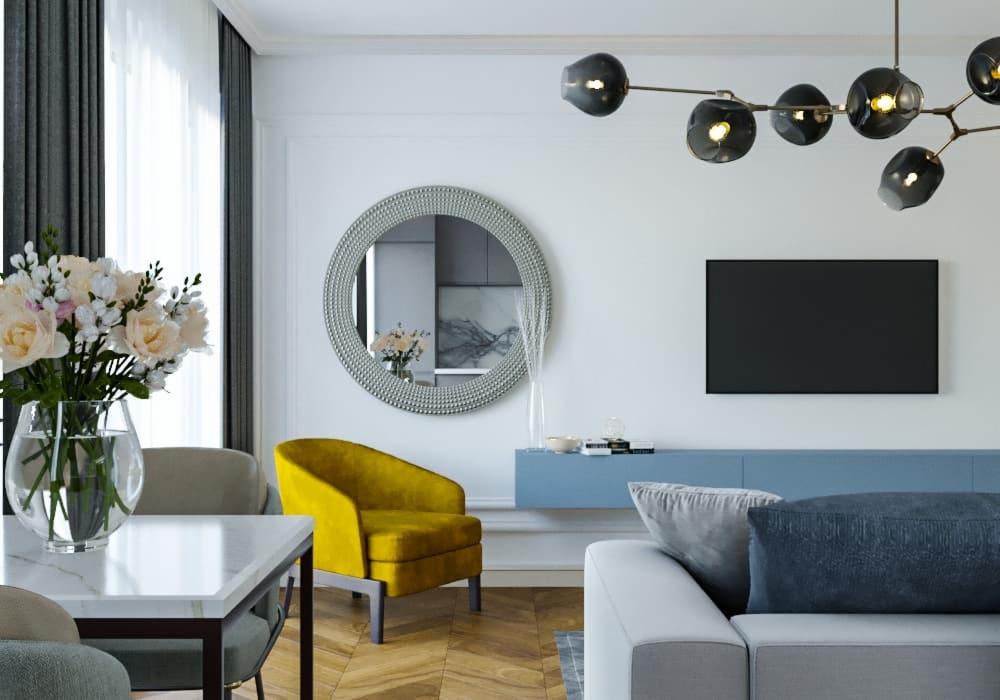 Jasne wnętrze salonu z parkietem, żółtym fotelem, szarą sofą, okrągłym lustrem w ozdobnej ramie i kwiatami w wazonie na stole z marmurowym blatem.