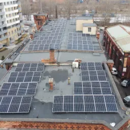 Widok z góry na dużą instalację fotowoltaiczną na dachu budynku w otoczeniu zabudowy miejskiej, z widocznymi panelami słonecznymi ułożonymi w rzędach i kolumnach, kominami oraz zaparkowanymi...