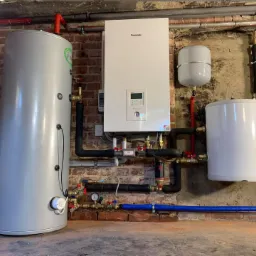Instalacja grzewcza z pompą ciepła Panasonic, zbiornikiem buforowym i naczyniem wzbiorczym, zamontowana na ceglanej ścianie. Widoczne liczne rury miedziane i zawory.