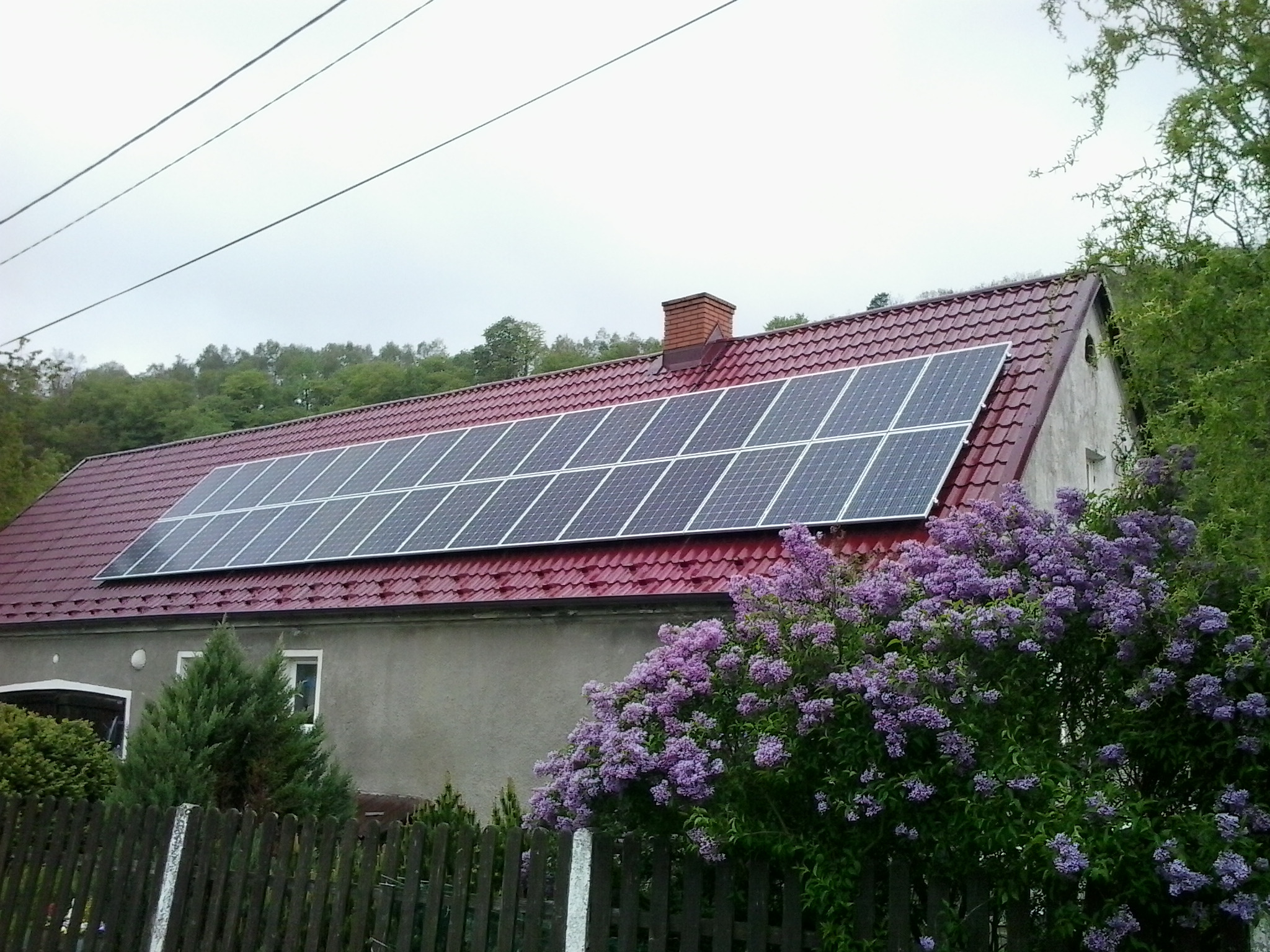 Dom jednorodzinny z panelami fotowoltaicznymi na czerwonym dachu, widoczne kwitnące bzy oraz drewniany płot w pochmurny dzień.