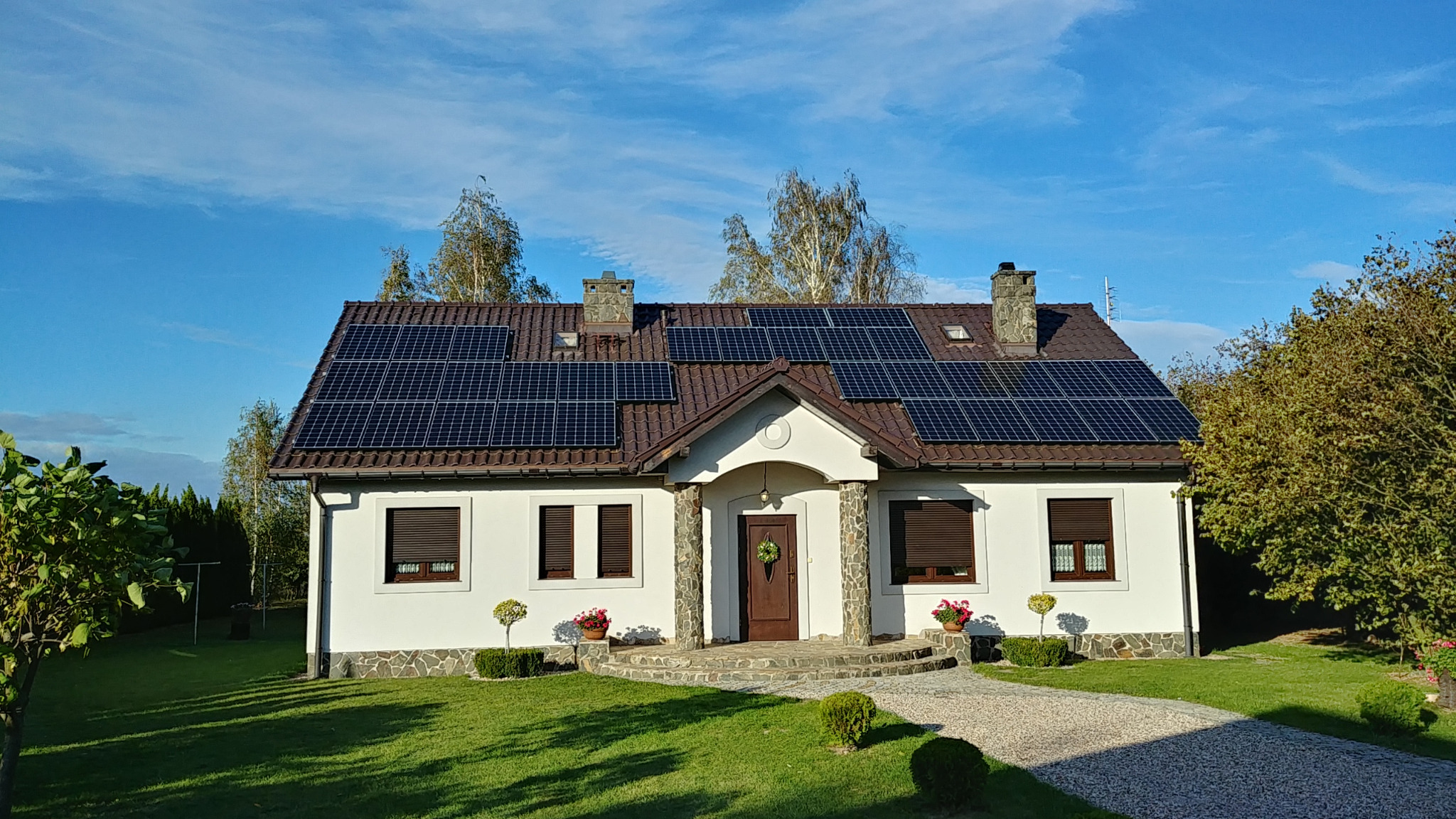 Biały dom jednorodzinny z brązowym dachem pokrytym panelami słonecznymi, widoczne kominy i zielony trawnik z ozdobnymi krzewami.