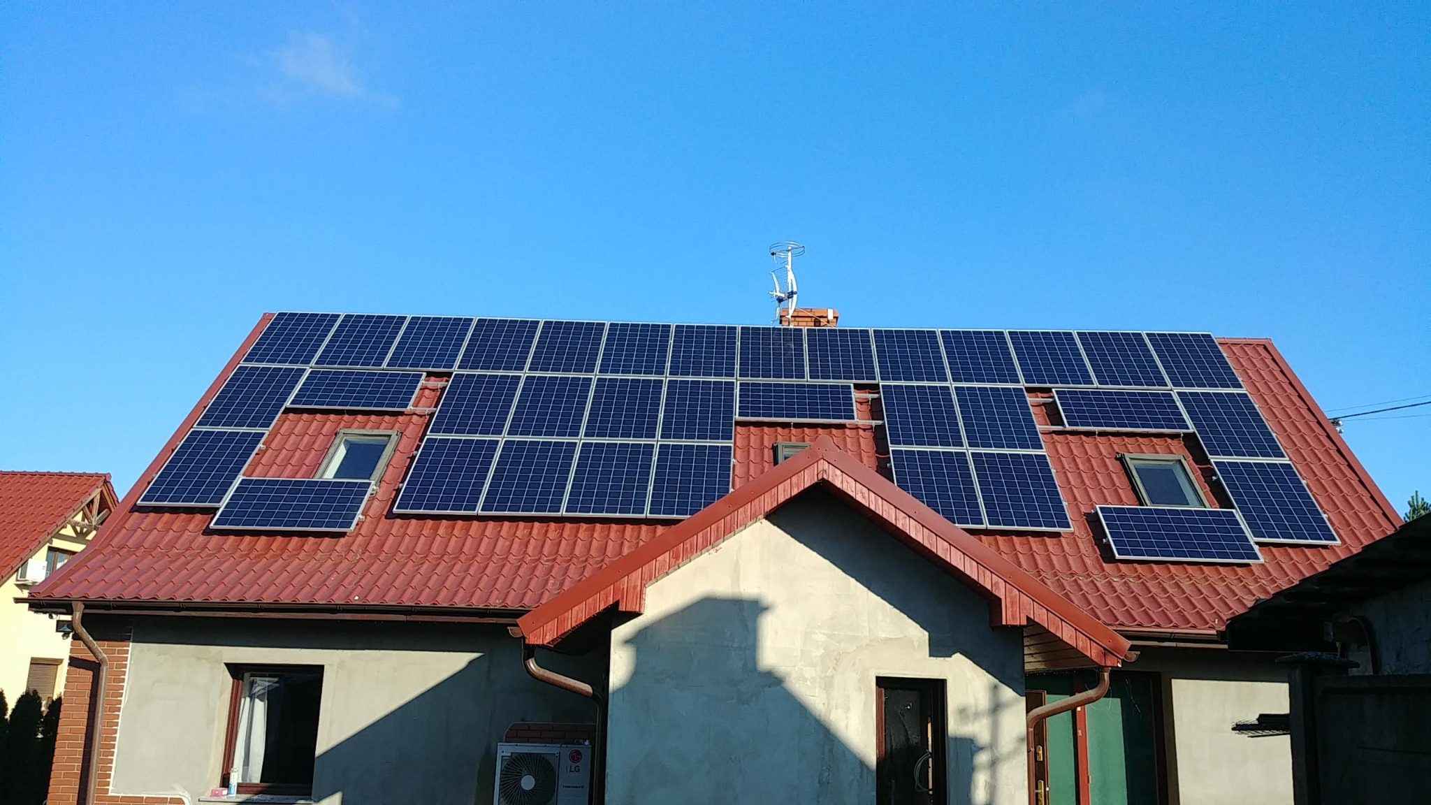 Czerwony dach domu jednorodzinnego z zamontowanymi panelami słonecznymi, okna dachowe wkomponowane w instalację, antena telewizyjna na kominie, błękitne niebo w tle.