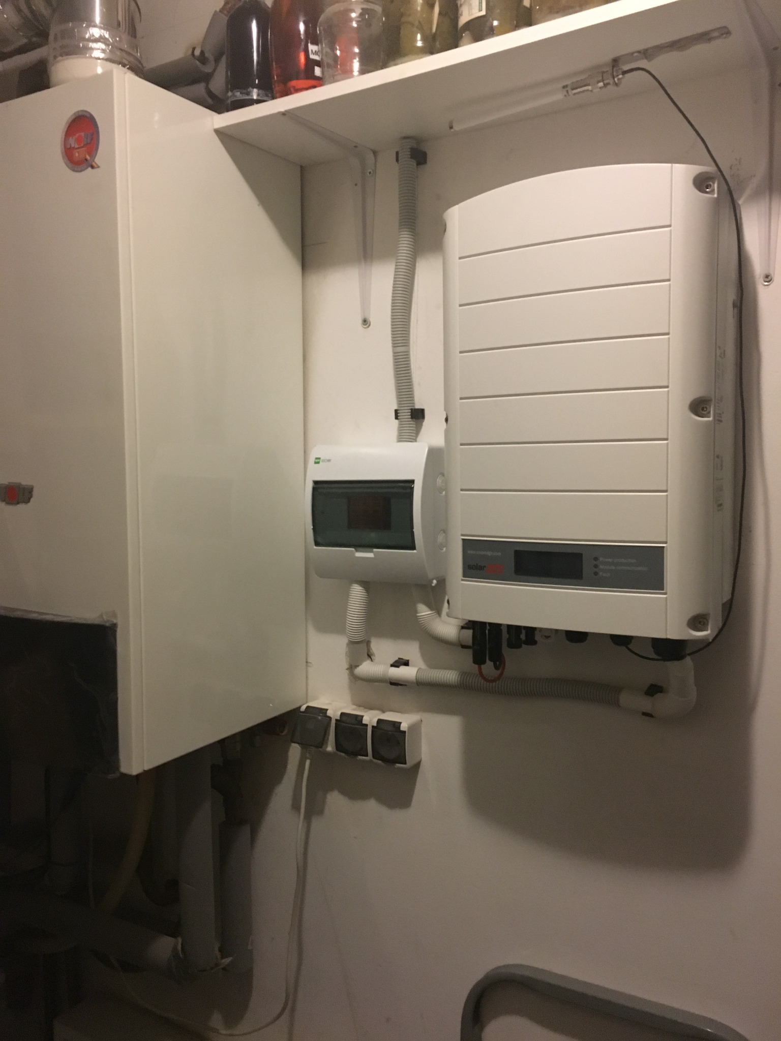 Biały inwerter fotowoltaiczny zamontowany na ścianie obok skrzynki elektrycznej i rur, z widocznymi przewodami i elementami instalacji w pomieszczeniu technicznym.