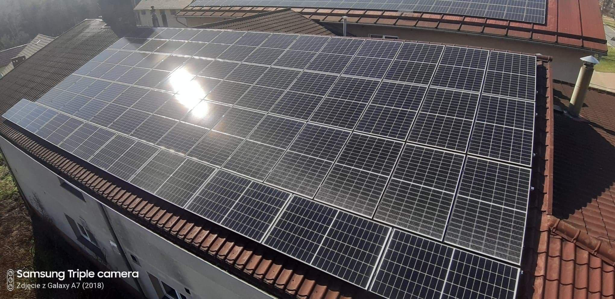 Instalacja paneli słonecznych na dachu domu z czerwonej dachówki, widoczne odbicie słońca na powierzchni paneli.