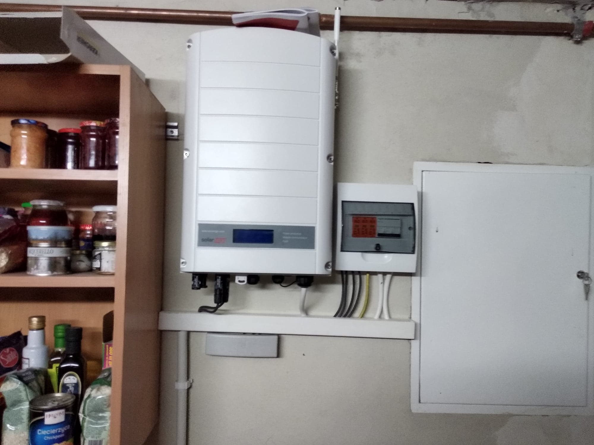 Biały inwerter solarny marki SolarEdge zamontowany na ścianie obok skrzynki elektrycznej, z widocznymi przewodami i elementami instalacji, częściowo zasłonięty przez półkę z zapasami spożywczymi.