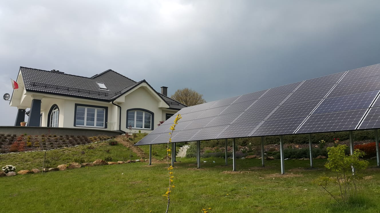Instalacja fotowoltaiczna na trawniku przed domem z flagą Polski, widoczna pochmurna pogoda.