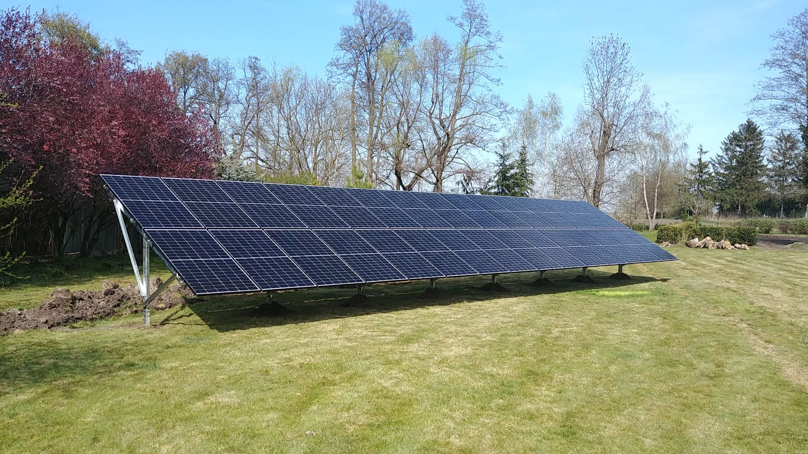 Długa instalacja paneli słonecznych na zielonym trawniku z drzewami w tle, widoczne ślady po pracach ziemnych.