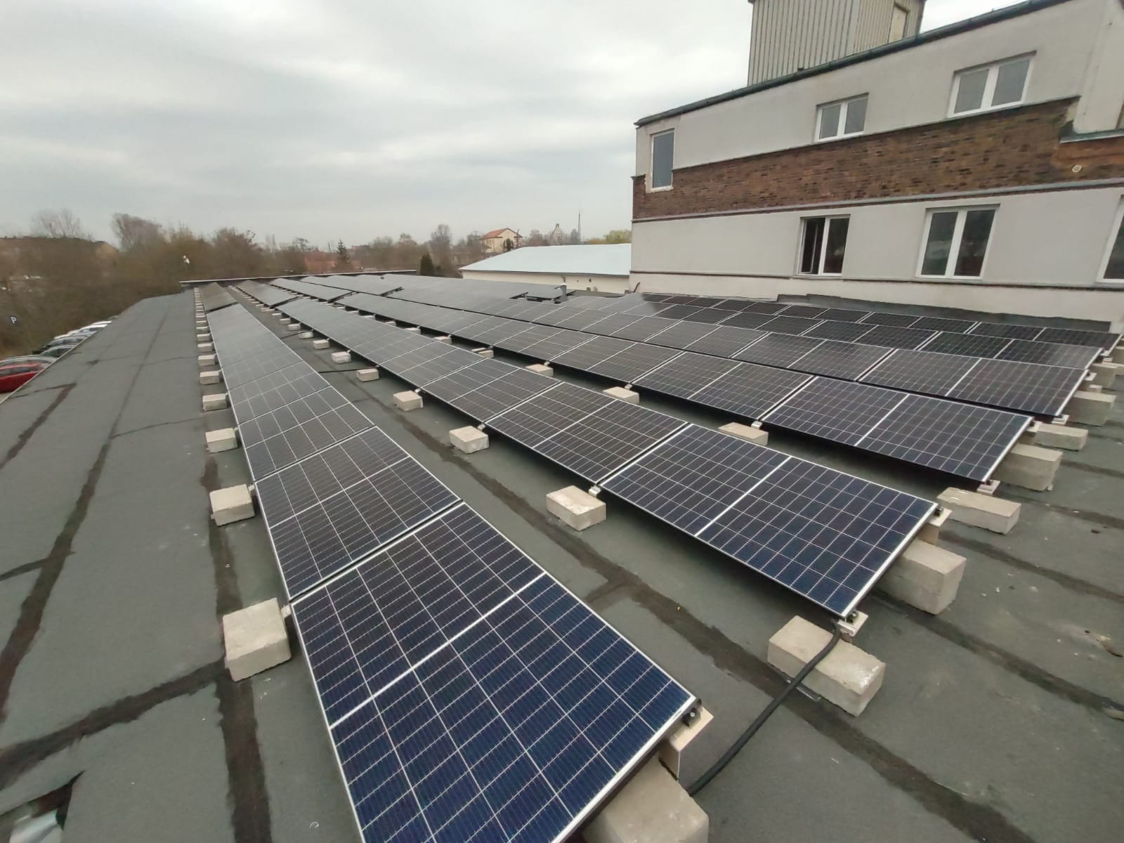 Rząd paneli słonecznych zamontowanych na płaskim dachu budynku przemysłowego, widoczne betonowe podstawy, pochmurne niebo w tle.