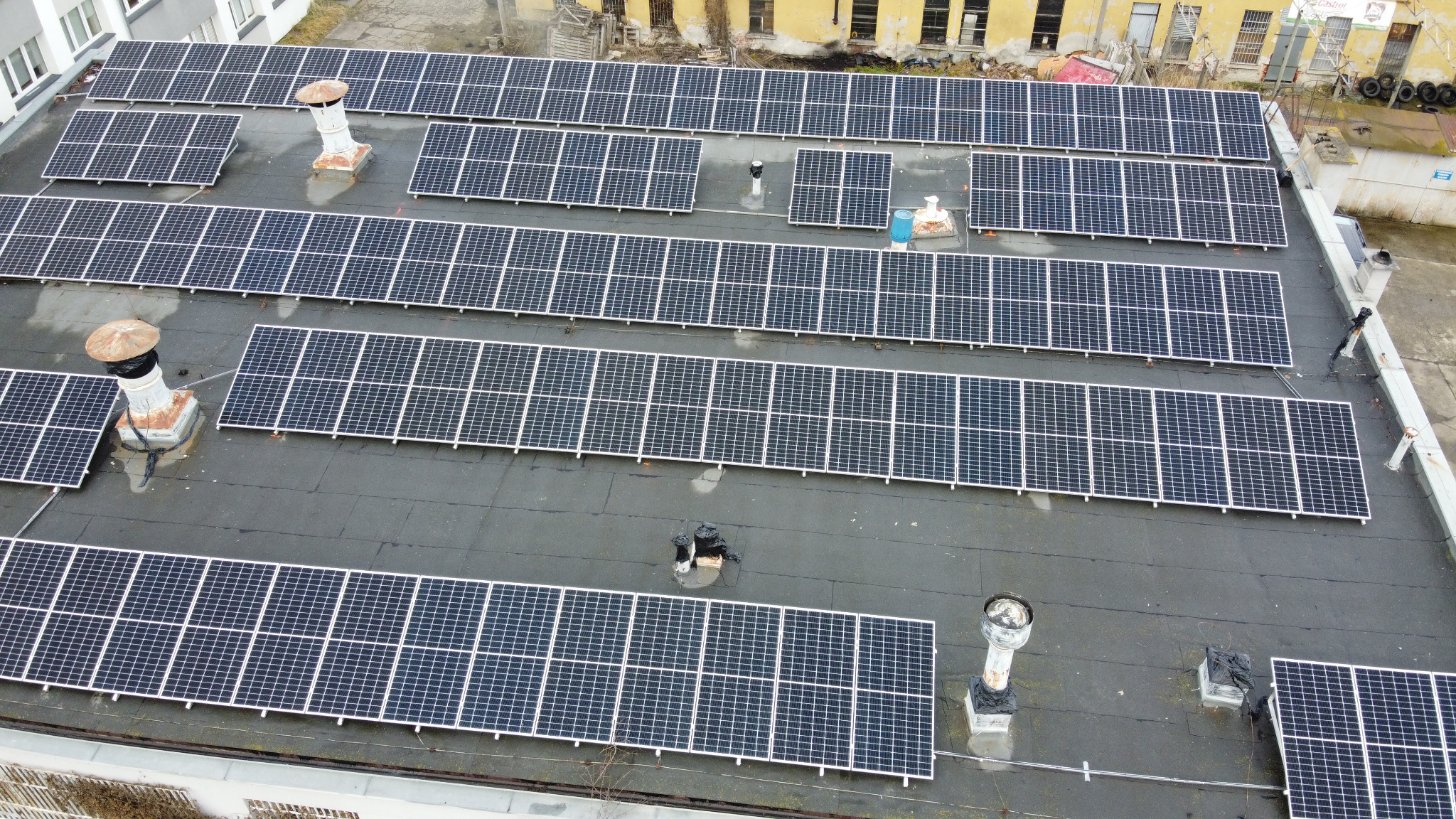 Widok z góry na liczne panele słoneczne zamontowane na płaskim dachu budynku, z kominami wentylacyjnymi i widocznymi sąsiednimi budynkami w tle.