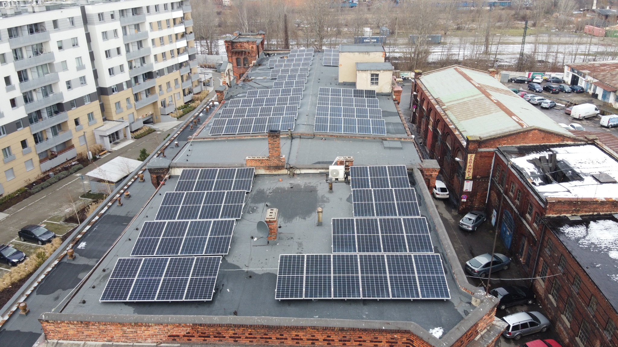 Widok z góry na dużą instalację fotowoltaiczną na dachu budynku w otoczeniu zabudowy miejskiej, z widocznymi panelami słonecznymi ułożonymi w rzędach i kolumnach, kominami oraz zaparkowanymi...