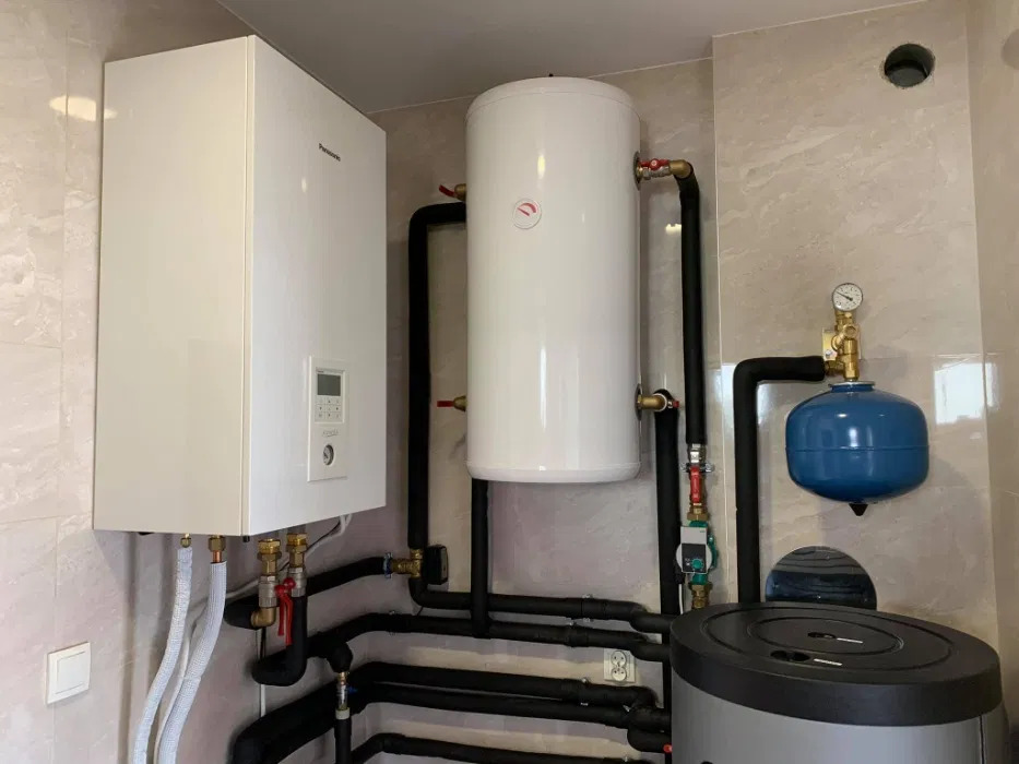 Zestaw urządzeń grzewczych w pomieszczeniu: biały wiszący bojler z wyświetlaczem, pionowy biały zbiornik akumulacyjny, niebieskie naczynie przeponowe i szary zasobnik ciepłej wody, połączone...