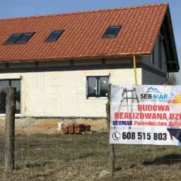 Niedokończony dom jednorodzinny z czerwonym dachem i oknami dachowymi, widoczny baner reklamowy firmy budowlanej przed posesją, na trawie stos dachówek.