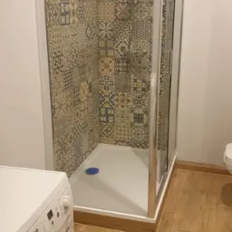 Wykończona łazienka z kabiną prysznicową wyłożoną mozaikowymi płytkami, widoczna pralka Bosch i drewnopodobna podłoga.