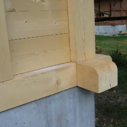 Fragment konstrukcji domu z jasnego drewna, widoczny detal połączenia belek na fundamencie betonowym, w tle trawnik i częściowo widoczny budynek.