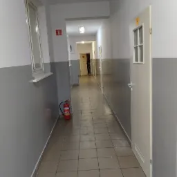 Długi korytarz biurowy z szarymi ścianami, brązowymi płytkami podłogowymi i czerwonym znakiem pożarowym, z gaśnicą przy ścianie, widok w głąb korytarza z otwartymi drzwiami na końcu.
