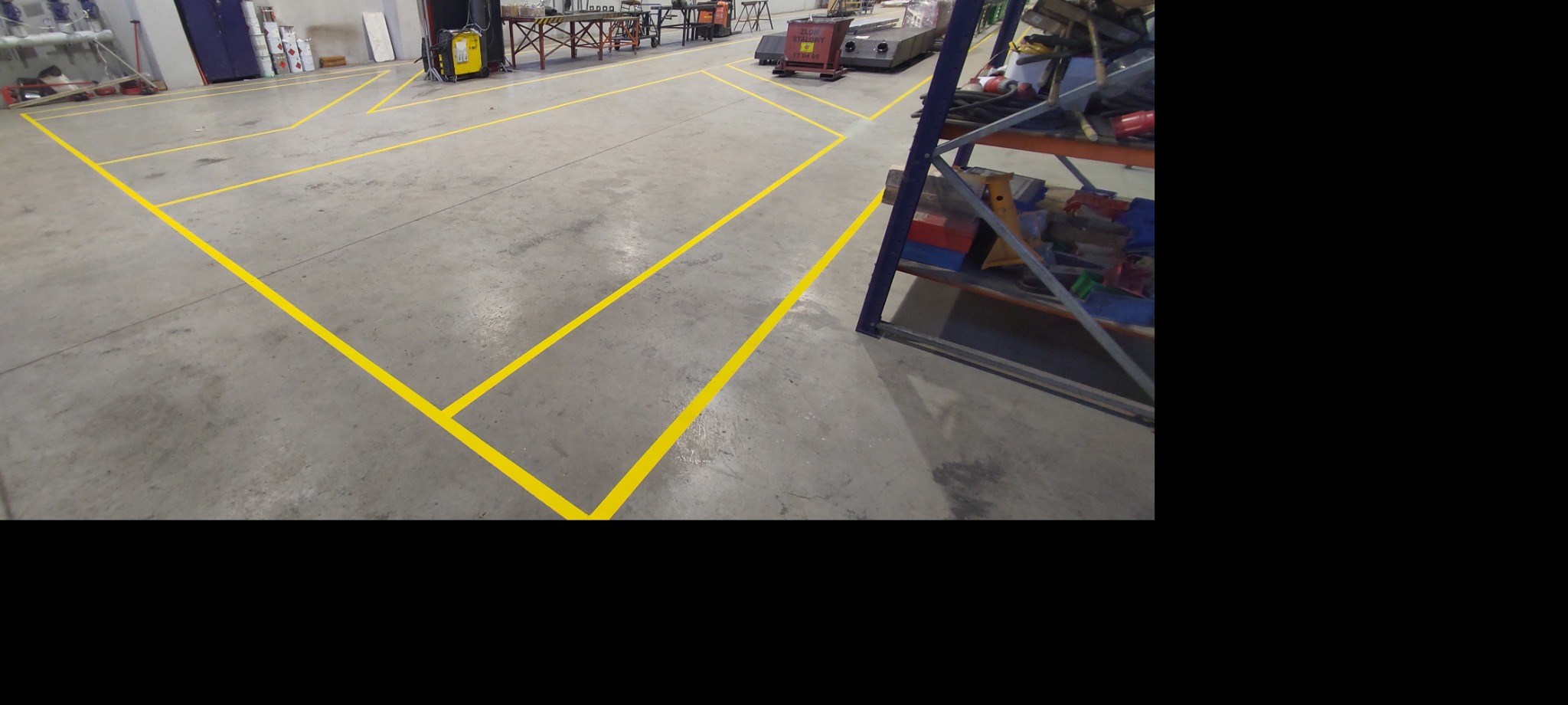 Żółte linie wyznaczające strefy na betonowej podłodze hali przemysłowej, w tle regał magazynowy i sprzęt spawalniczy.