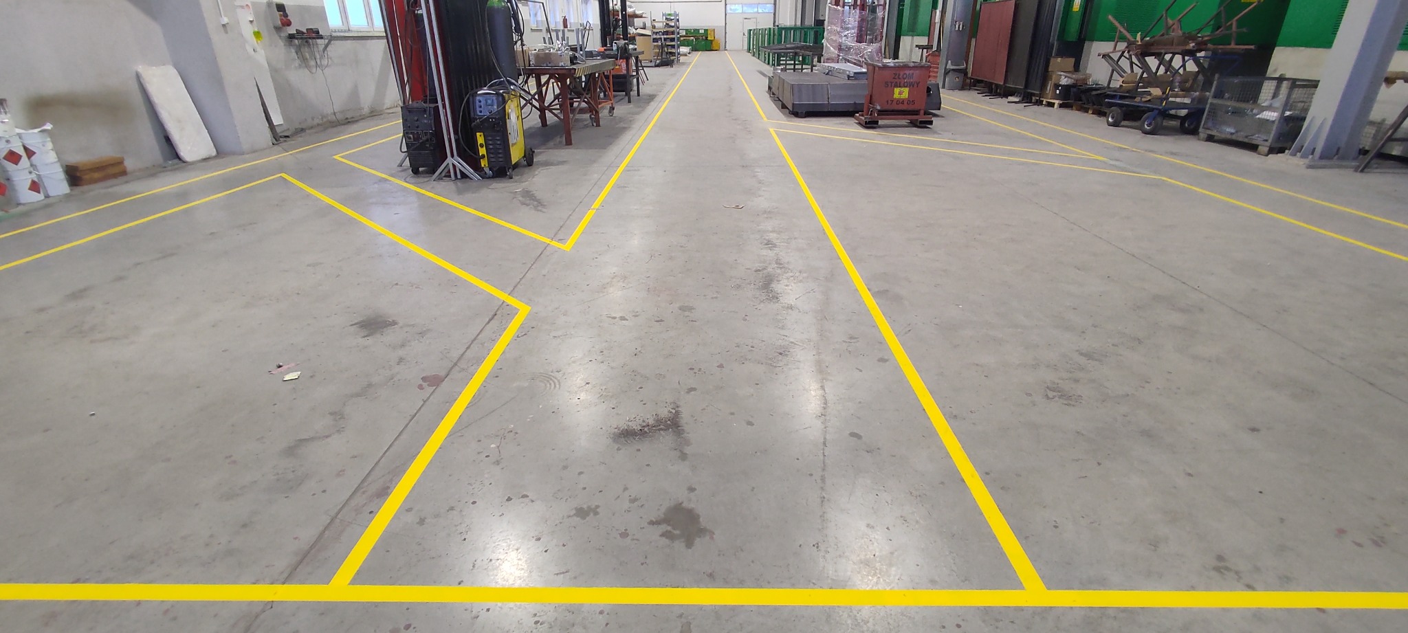 Szary betonowy magazyn z wyznaczonymi żółtymi liniami komunikacyjnymi. Widoczne wózki, regały i sprzęt spawalniczy.