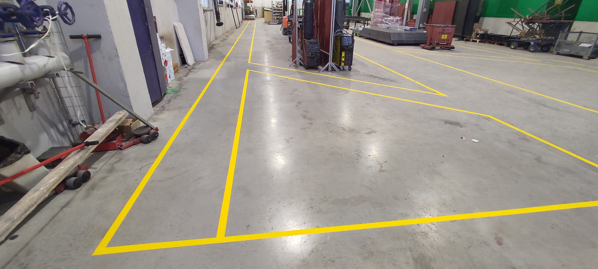 Żółte linie wyznaczające strefy na posadzce hali przemysłowej, widoczne wózki widłowe i inne maszyny w tle, posadzka betonowa, oświetlenie sufitowe.