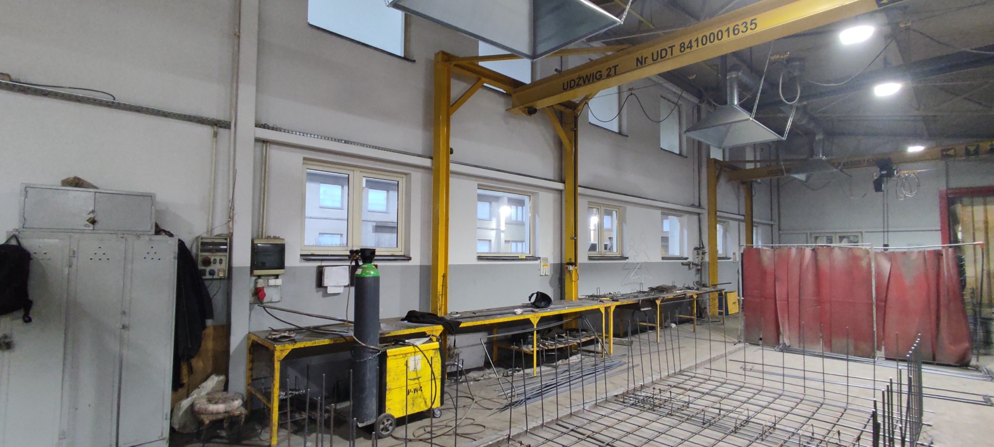 Wnętrze hali produkcyjnej z suwnicą Udzwig 2T, stołami roboczymi z butlą gazową i przygotowanymi elementami zbrojeniowymi do spawania, widoczne okna i osłona spawalnicza.