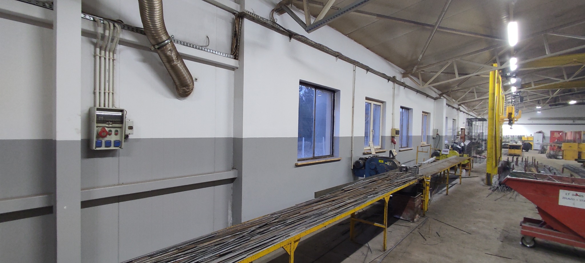 Wnętrze hali produkcyjnej z szarymi ścianami do połowy wysokości, żółtym stołem roboczym z ułożonymi prętami, widoczne okna, instalacje elektryczne, suwnica i pojemnik na odpady.