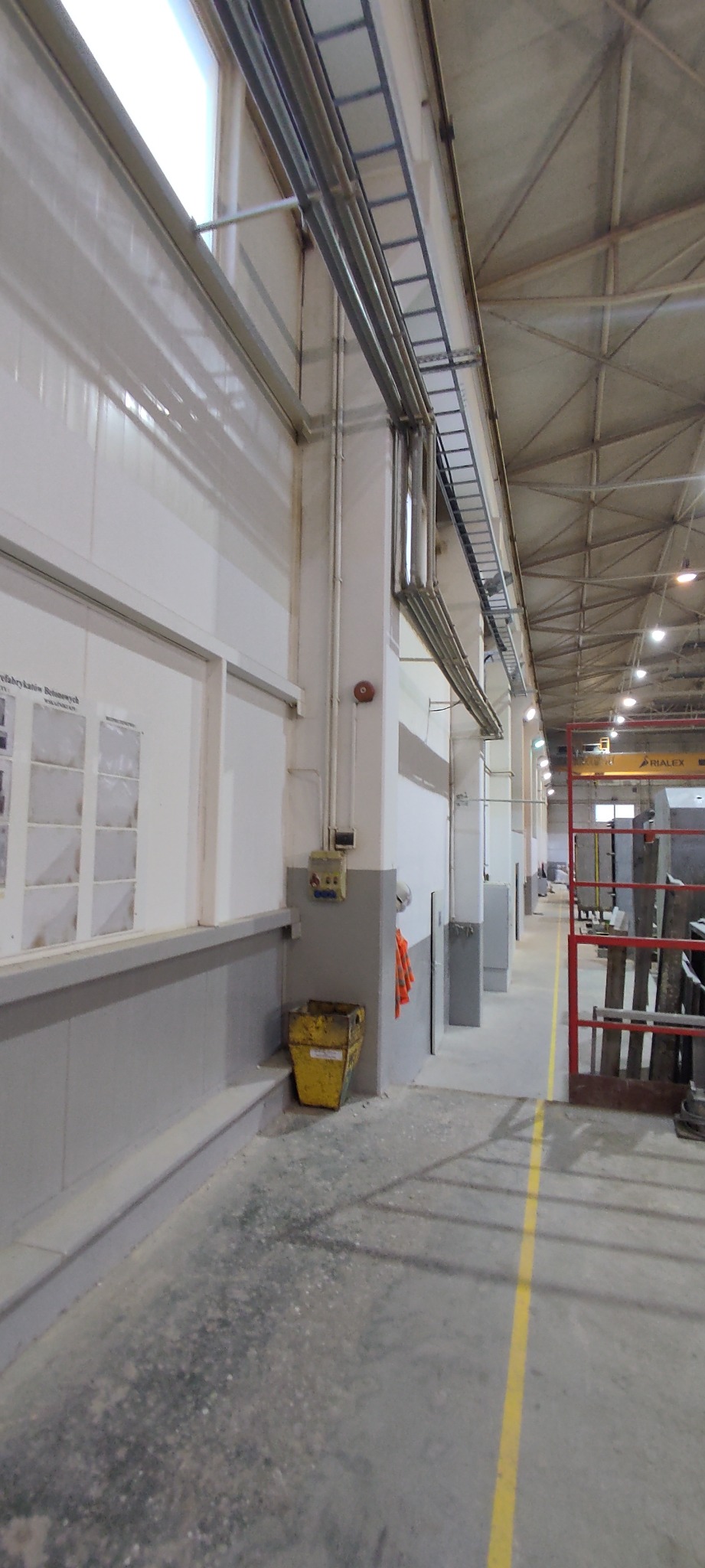 Wnętrze hali przemysłowej z widocznymi instalacjami pod sufitem, ścianami pomalowanymi do połowy na szaro, żółtym koszem na śmieci przy ścianie i żółtą linią na podłodze wyznaczającą ścieżkę.
