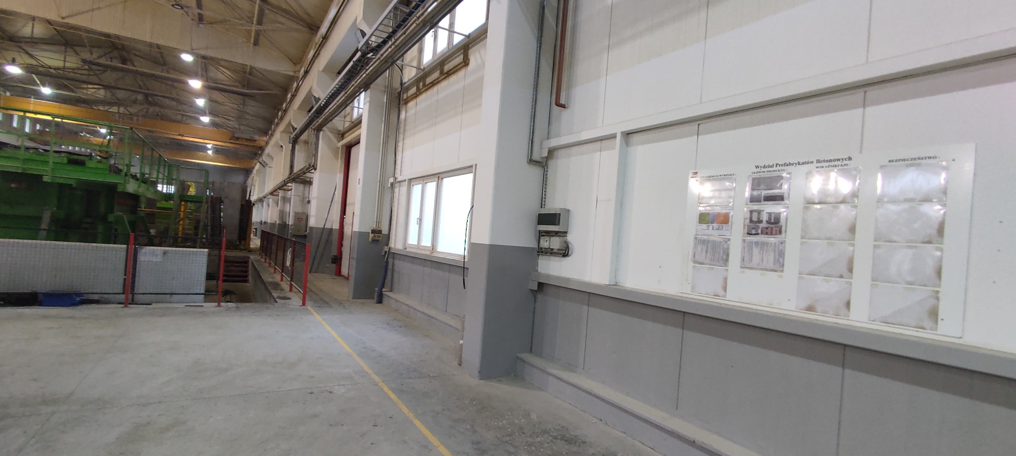 Wnętrze hali przemysłowej z betonową podłogą, widoczne maszyny, ściany z szarym cokołem i tablice informacyjne, oświetlenie jarzeniowe.