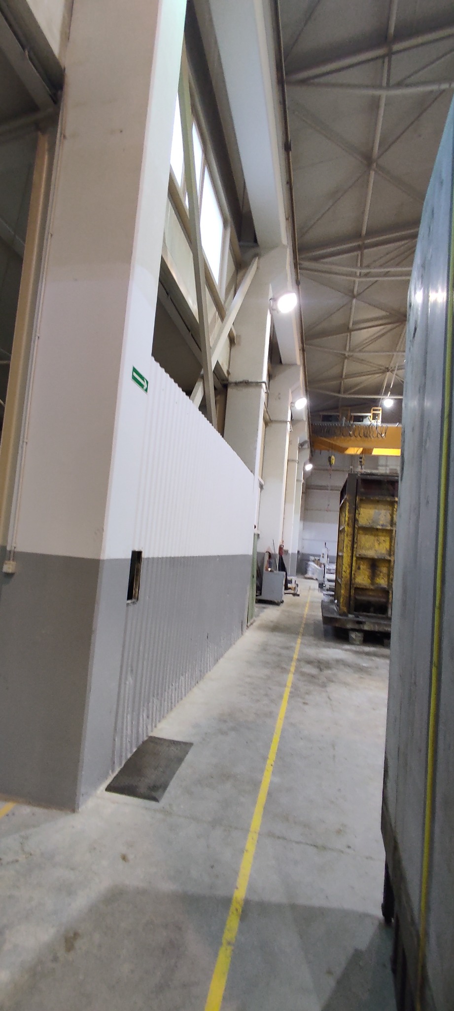 Wnętrze hali przemysłowej z betonową podłogą, żółtą linią wyznaczającą ścieżkę, widocznym fragmentem ściany z zieloną strzałką kierunkową i oknami w górnej części, oraz elementami konstrukcji dachu...