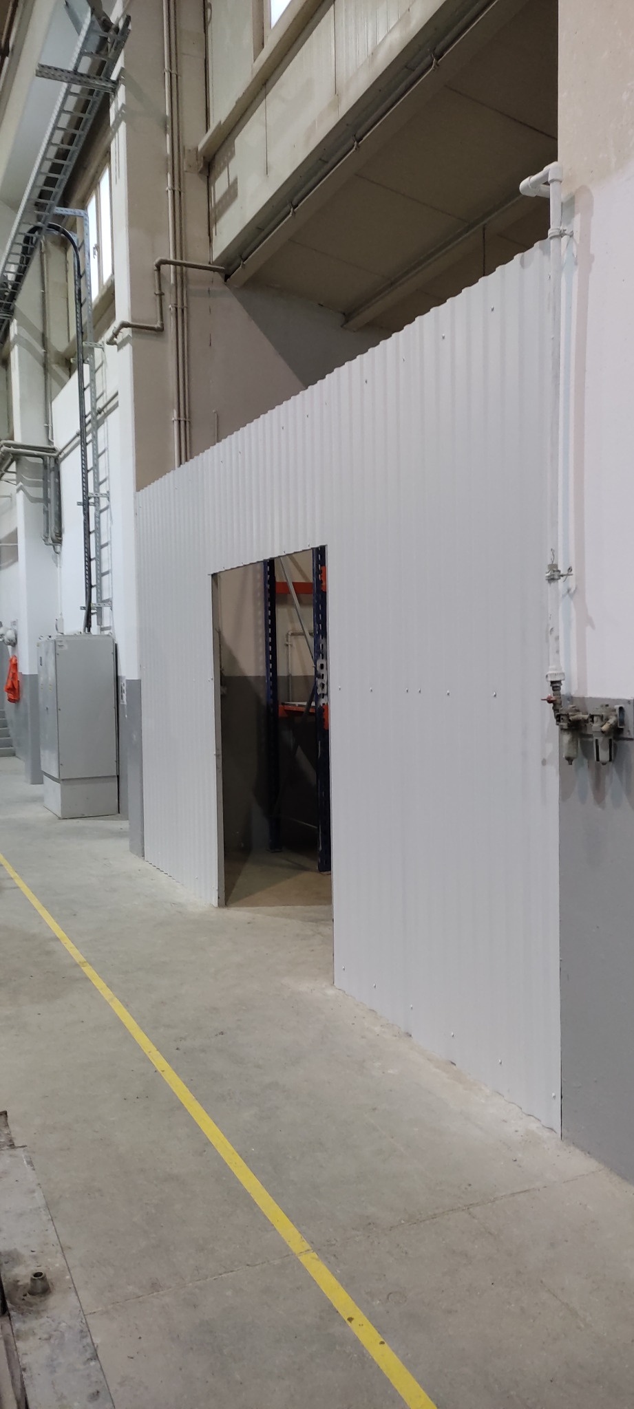 Wnętrze hali produkcyjnej z nowo postawioną, białą, falistą ścianą działową z otworem drzwiowym, widoczne instalacje pod sufitem i żółta linia na posadzce.