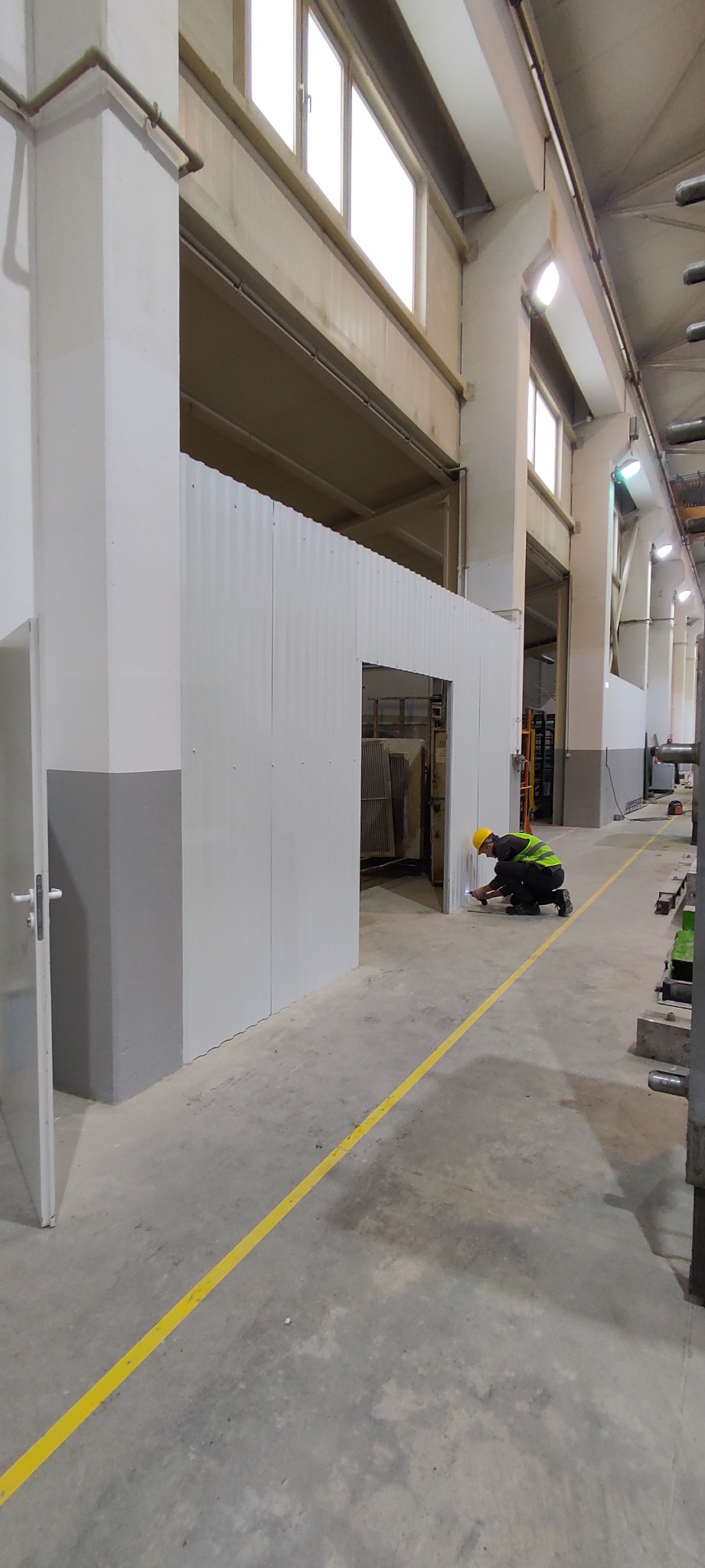 Pracownik w kasku i kamizelce odblaskowej uszczelnia dolną krawędź białej ściany działowej w hali przemysłowej, z widoczną żółtą linią na posadzce.