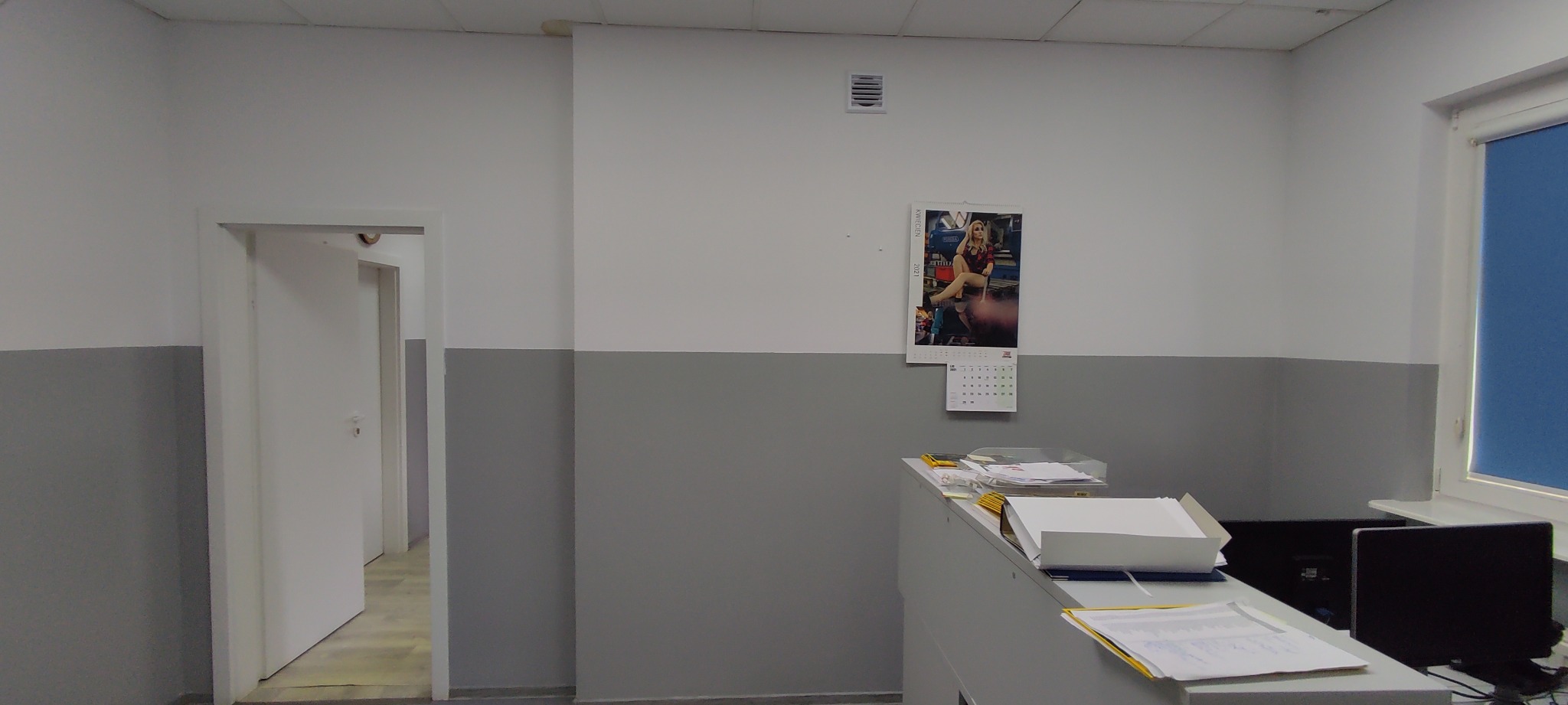 Wnętrze biura po malowaniu: ściany pomalowane na biało i szaro, widoczny fragment biurka z dokumentami, kalendarz na ścianie, drzwi do innego pomieszczenia.