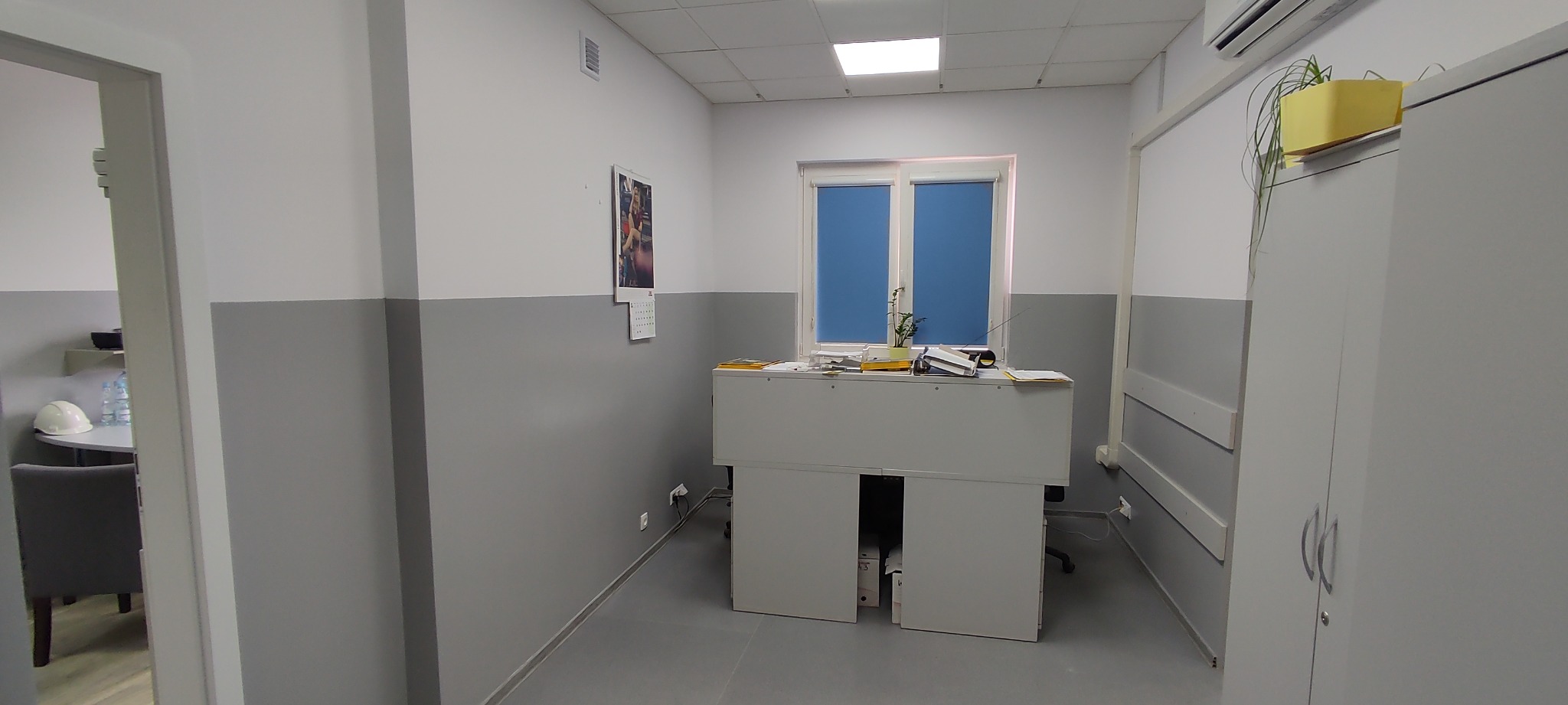 Biuro z dwukolorowymi ścianami, białymi meblami, biurkiem z dokumentami i rośliną w żółtej doniczce na szafce, widok z perspektywy wejścia.