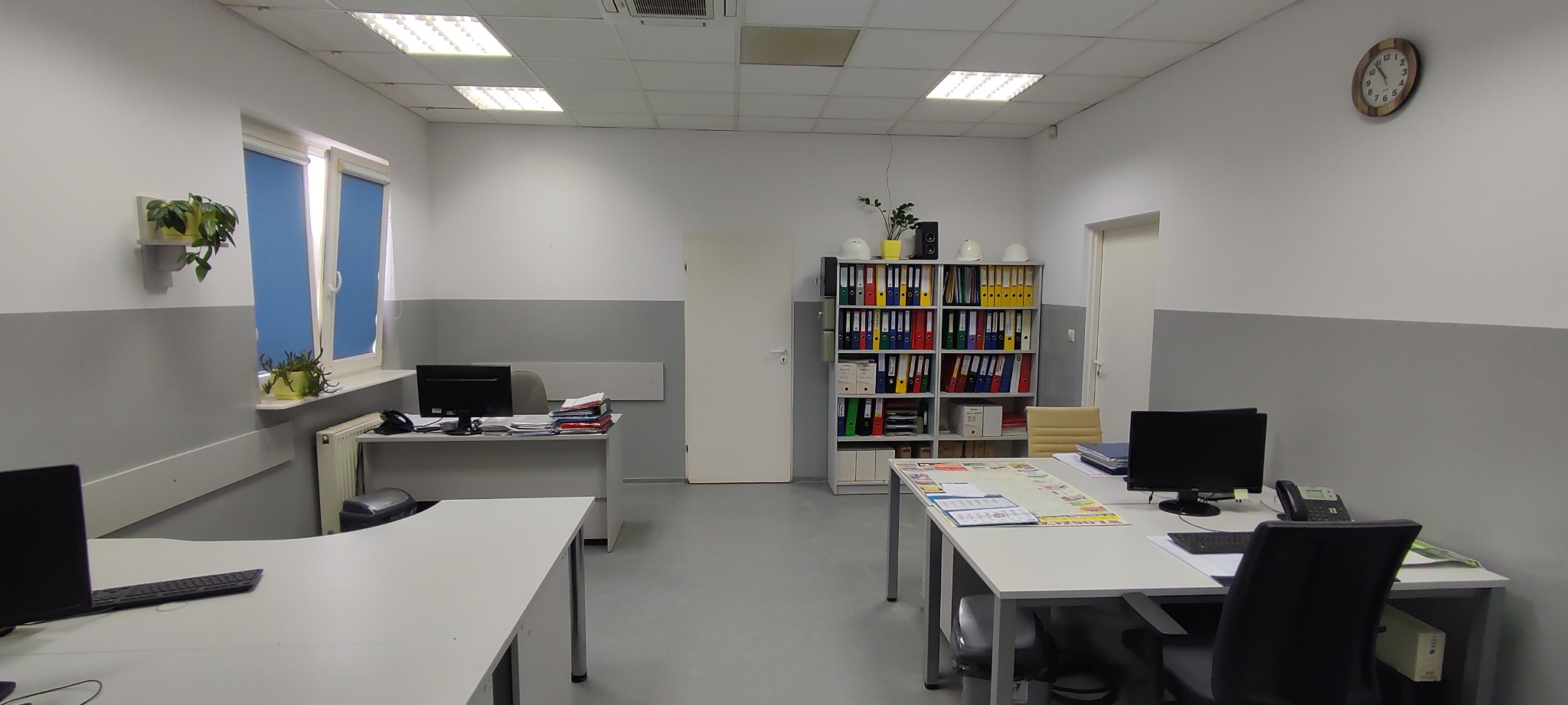 Jasne biuro z białymi ścianami i szarym pasem, biurka z komputerami, regał z segregatorami i kaskami ochronnymi, roślinki w doniczkach na parapecie i ścianie, zegar na ścianie.