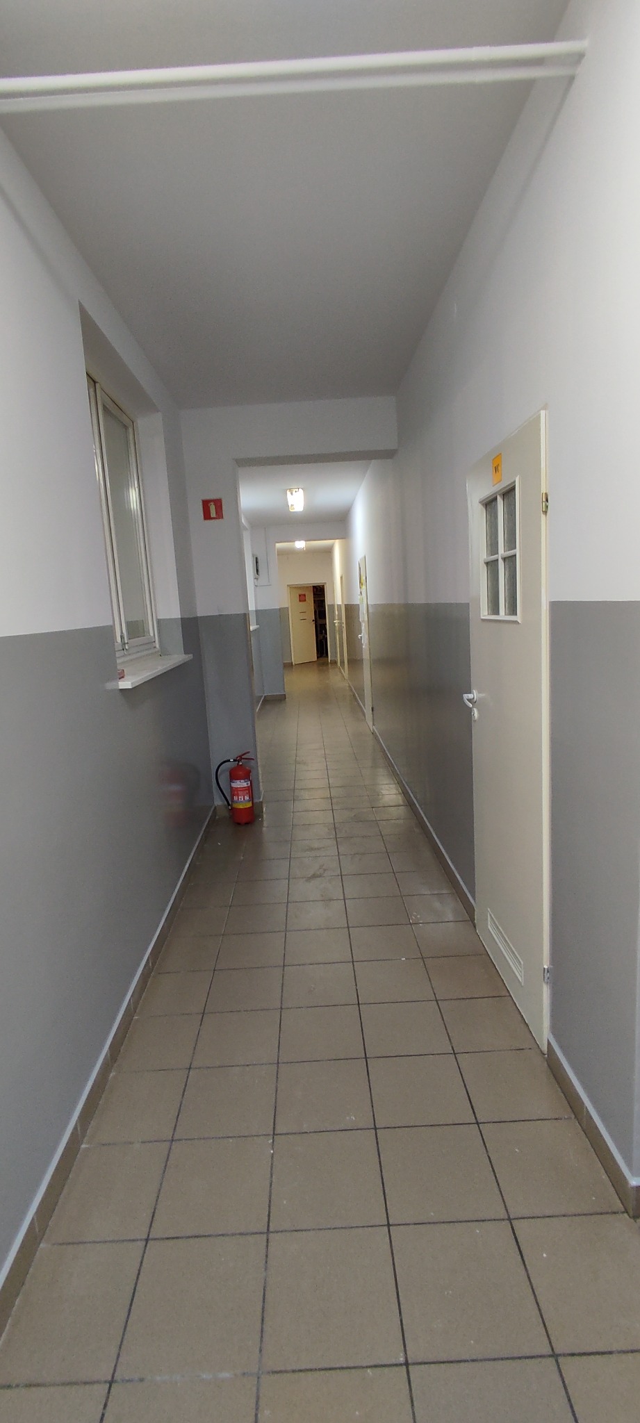 Długi korytarz biurowy z szarymi ścianami, brązowymi płytkami podłogowymi i czerwonym znakiem pożarowym, z gaśnicą przy ścianie, widok w głąb korytarza z otwartymi drzwiami na końcu.